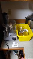 Cuisinart motor base with attached transparent work bowl and feed tube, power cord visible, unit sitting on kitchen counter next to a yellow container.
