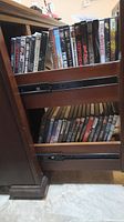 Photo shows two pull-out shelves filled with assorted DVDs including titles like The Hobbit, Clash of the Titans, The Expendables, Sin City, and others.