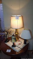Photo showing four assorted table lamps and a lit tree on a round wooden table. The brass lamp with cream shade is lit, the smaller white shade lamp and floral glass shade lamp are unlit, and the lit tree emits soft light.