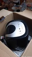 Air fryer partially inside open cardboard box, showing black and silver plastic exterior and handle.