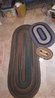 Photo of three braided area rugs of varying sizes on a floor, showing largest rug as an elongated oval multicolor design, medium-sized rug with bear and pinecone motif, and small navy and cream oval rug.