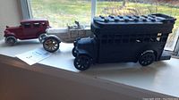 Side view of three cast iron cars displayed on a window sill: black school bus, white convertible with dog figure, and red vintage car.