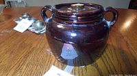 Front side view of vintage brown stone glazed bean pot with lid and two handles sitting on a wooden table.