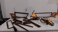 Photo showing three wooden-handled hacksaws, hand drills, and other antique metal tools arranged on a table.