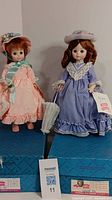 Two Madame Alexander dolls standing on a box, one in a purple dress and the other in a peach dress, both holding umbrellas.