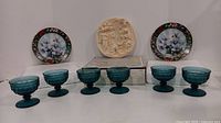 Wide view showing two hummingbird collector plates, six blue glass compotes, and a beige decorative plaque on a box.