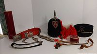 Full display of all items in the lot arranged on a surface including hats, belt buckles, lanyards, red cylindrical container, leather strap, and wooden artifact.