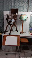 Vintage wooden high chair, floor globe, and drafting table shown together on white background with wallpaper behind