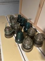 Ten vintage glass electrical insulators arranged on a wooden surface, showing variation in color from clear to dark green and different styles, some wire-bound.