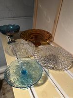 Five vintage glass serving pieces including cake stands and serving plates on display shelf under lighting