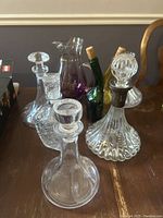 Photo showing assorted clear glass decanters and two smaller green and amber glass bottles with cork stoppers, alongside a purple-tinted glass pitcher.