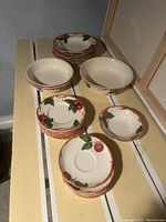 Assorted plates and bowls made by Franciscan, showing floral and leaf pattern in green and reddish-orange on cream background, arranged on wooden surface.