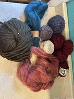 Photo of assorted yarn showing large black bulky yarn skein, blue and burgundy intertwined yarns, pink fluffy fiber, and smaller balls in purple and beige.