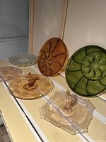 Photo showing six assorted vintage glass serving platters and trays in amber, green, and clear glass with various decorative patterns, arranged on a wooden surface.