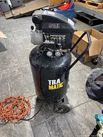 Front angle of vertical black air compressor showing handle, wheels, orange extension cord, and TRA MATIC branding.