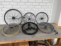 Six bicycle rims displayed on a table against a white brick wall, showing various sizes and models including metal spoked rims and a three-spoke black rim.
