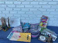 Overview of various packaged party supplies laid out on a blue cloth with a white brick wall background. Includes balloon column kit, bubble solution, party favors, and packaged napkins and plates.