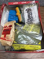 Top-down view of box containing mixed train hobby items including bags of gravel and blended turf, yellow control unit, and plastic fence pieces.