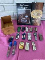 Photo showing three cigar books, wooden cigar box without lid and multiple assorted lighters and two bottles of butane fuel.
