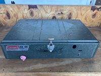 Top view of closed green metal Coleman stove case with carrying handle and latch on front, showing some wear and dust.