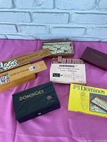 Photo of multiple domino game sets displayed on pink cloth, showing variety of packaging and boxes including wooden and cardboard ones