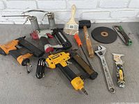 Overview of the entire lot showing all items laid out on a gray surface against a white brick background. Includes pneumatic nailers, oil cans, mallet, paintbrush, wrench, screwdriver, utility knife.
