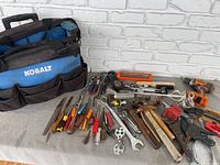 Full view of handyman tools spread in front of the Kobalt wheeled carrying case including hammers, screwdrivers, pliers, wrenches, sockets, clamps, pneumatic cutting tool, and cordless screwdriver without battery.