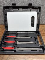 Top view of the black plastic carrying case holding four utility knives and a white plastic cutting board.