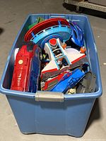 Large blue plastic bin filled with assorted toys including a red and blue bus-like vehicle, blue playset ramps, and a blue helmet.