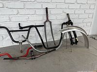 Photo showing multiple bicycle parts including black and silver handlebars, grey front fork with red paint spots, black suspension fork with rust, chrome fender, and red rod.