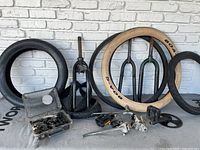 Photo showing tires, front forks, kickstands, pedals, sprockets, and plastic box with small parts on carpet against white brick wall.