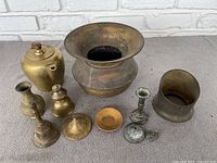 Lot of various brass items arranged on a grey surface including an old style spittoon, teapot, multiple vases, a candlestick holder, and more.
