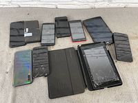 Lot of 10 assorted smartphones and tablets laid out on a carpet, showing devices with cracked screens and a Samsung phone with reflective back