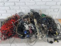 Photo showing a large pile of assorted cables including power cords, speaker cords, and various computer wires tangled and piled on a surface in front of a white brick wall.