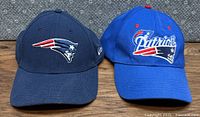 Front view of two New England Patriots ball caps, one navy blue with team logo and one royal blue with team script logo.
