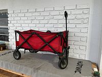 Red fold-up wagon showing side profile with fabric storage area, black metal frame, and wheels on outdoor surface.