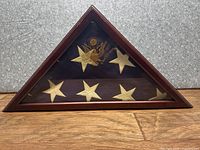 Folded American flag in a triangular wooden display case, showing gold embossed eagle emblem and stars on blue field.