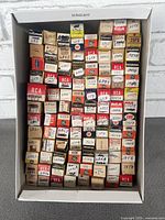 Box with vintage vacuum tubes stacked tightly, showing assorted brands and handwritten labels of model numbers.