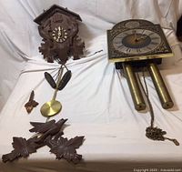 Two vintage clocks together: one brass and metal Tempus Fugit wall clock with two brass weights, and one wooden cuckoo clock with detached parts arranged on white background.