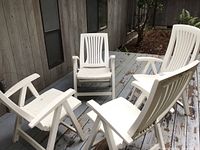 Four white plastic Adirondack chairs arranged on a painted wood deck, showing front views, backs, and armrests. Chairs are foldable with slatted designs.