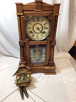 Front view showing both clocks including the taller wooden wall clock with columns and smaller Swiss cuckoo clock with pinecone weights.