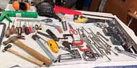 Wide view showing assortment of hand tools laid out on surface, including hammers, screwdrivers, socket wrenches, clamps, staplers, tape measure, and safety equipment.