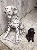 Front and side view of large Dalmatian dog statue and small black dog statue on cement ground with wooden wall background.