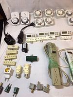 Overview of mechanical dial timers, two digital timers, various plug adapters, outlet adapters with light sockets, an off-white power strip with multiple outlets, and additional small plug connectors.