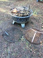 Fire pit set outdoors on grass with fire started using sticks inside the fire pit. Lid and cooking grate shown on ground.
