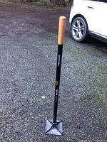 Full view of Fiskars heavy duty tamper standing upright on gravel, showing orange grip and square steel head.