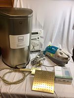 Photo showing stainless steel step trash can with foot pedal, ironing board cover packaging, Dirt Devil spot scrubber vacuum, metal perforated plate, and cords.