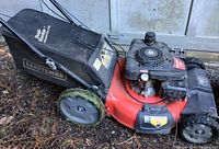 Red Craftsman lawnmower with black Dusk Buster grass bag attached, sitting on dirt floor with some grass on wheels.