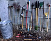 Photo of assorted long handled garden tools including rakes, hoes, shovels, cultivators, pruners and a galvanized metal trash can for storage, showing full lot from the side.