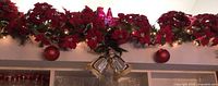Wide shot of red poinsettia garland with string lights and large red ornaments hung over a kitchen cabinet.
